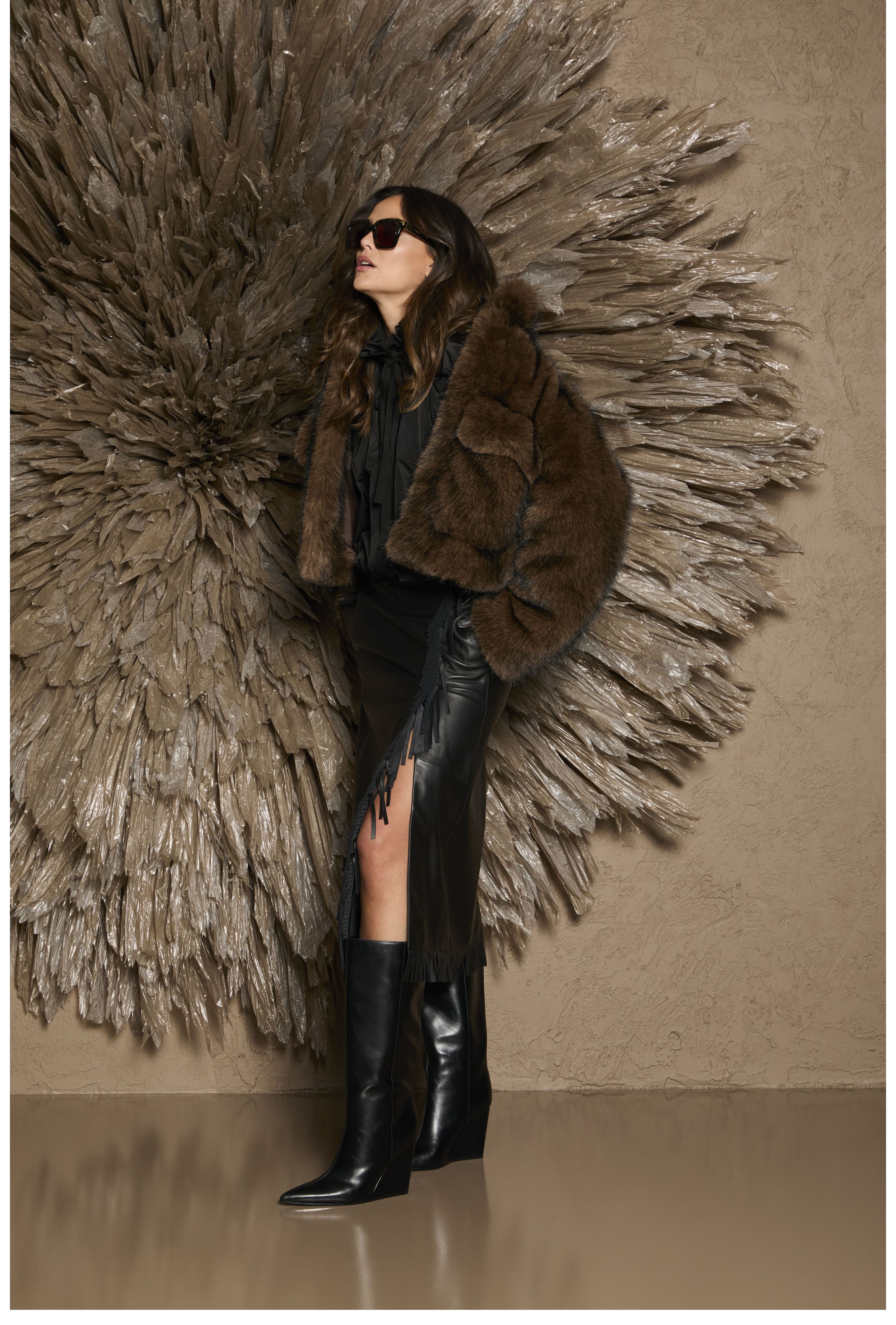 Stylische Person mit Pelzjacke, schwarzen Highlights und Stiefeln vor dekorativer Wand.