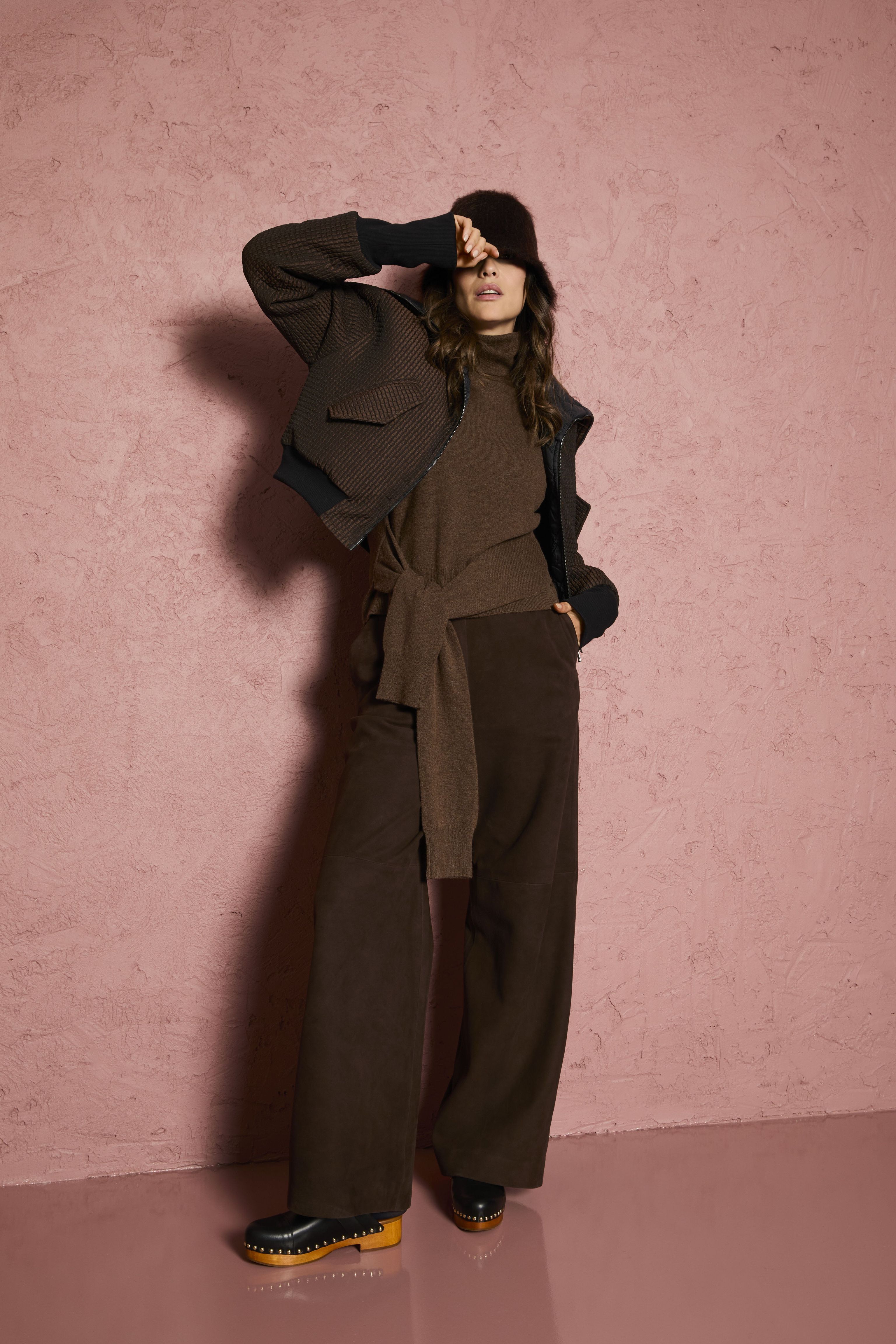 Frau in oversized Pullover, weiten Hosen und Stiefeln lehnt lässig an Wand, Sonnenbrille im Haar.