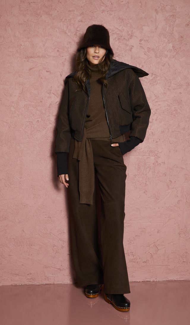 Frau trägt stylischen olivfarbenen Anzug mit Jacke, Schal und flachen Schuhen vor rosa Wand.