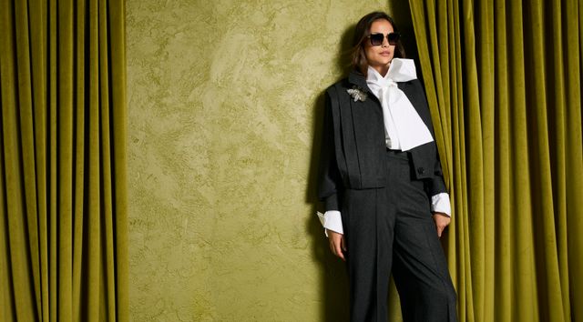 Person in elegantem Anzug mit Weste, weißem Hemd und Sonnenbrille vor grünem Vorhang.