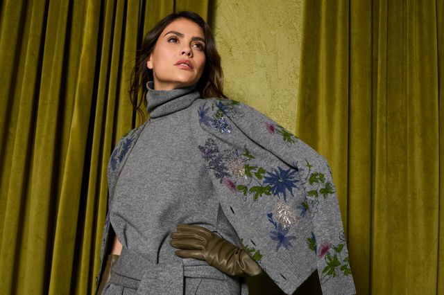 Frau trägt stylischen Tweedmantel mit floralem Schal vor gelbem Vorhang.