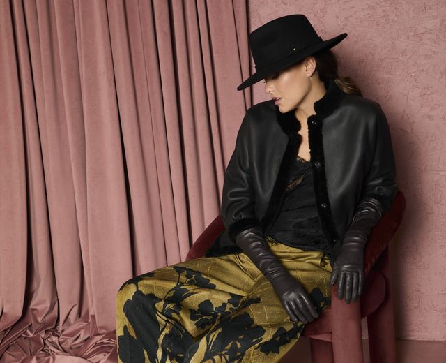 Stylische Frau in schwarzem Hut, Lederjacke und floral-goldenen Hosen sitzt elegant auf Stuhl.