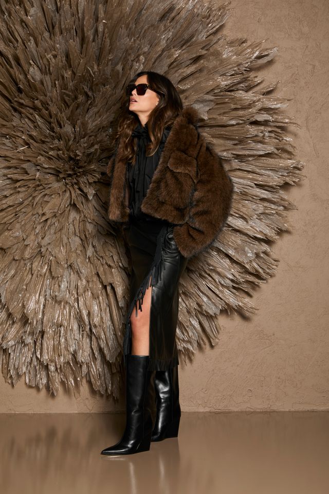 Stylische Person mit Pelzjacke, schwarzen Highlights und Stiefeln vor dekorativer Wand.